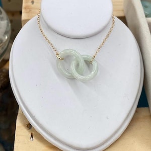 May include: A delicate gold chain necklace with a pendant featuring two interlocking jade rings. The rings are a pale green color and have a smooth, polished finish.