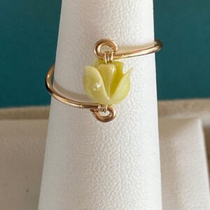 May include: A gold wire ring with a yellow flower charm. The ring is on a white display stand.