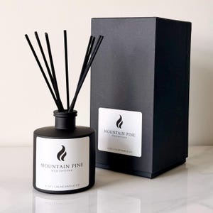 May include: A black reed diffuser with black reeds and a matching black box. The diffuser has a white label with the text "Mountain Pine Reed Diffuser" and a flame logo. The diffuser is 8 oz.