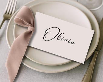 May include: A place setting with a white plate, a fork, and a knife. A light pink ribbon is tied in a bow and placed on the plate. A white card with the name "Olivia" in black script is on the plate.