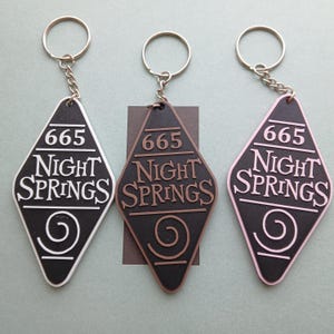 May include: Three keychains with a diamond-shaped tag that says "665 Night Springs" with a swirl design. The keychains are white, brown, and pink.