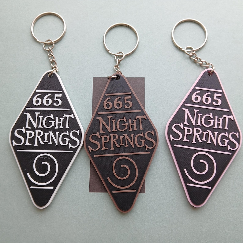 May include: Three keychains with a diamond-shaped tag that says "665 Night Springs" with a swirl design. The keychains are white, brown, and pink.
