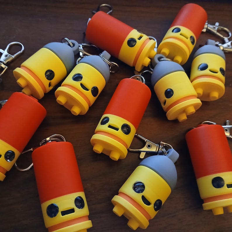 May include: A collection of novelty keychains with a cylindrical design. The keychains feature a red top, yellow midsection, and a gray cap. Each keychain has a cartoon face with black eyes and a smiling mouth. The keychains are attached to silver key rings.