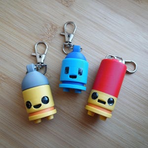 May include: Three colorful keychain charms in the shape of spray paint cans. One is yellow and gray, one is blue, and the other is red. Each has a silver clasp and a smiling face design. The keychains are on a wooden surface.