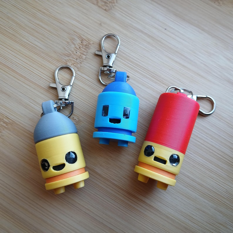 May include: Three colorful keychain charms in the shape of spray paint cans. One is yellow and gray, one is blue, and the other is red. Each has a silver clasp and a smiling face design. The keychains are on a wooden surface.