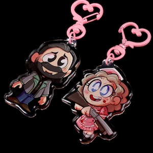 May include: Two cartoon character keyrings. One keyring features a man with a beard and a black jacket. The other keyring features a woman with blonde hair wearing a pink dress and holding a gun. Both keyrings have pink heart-shaped rings.