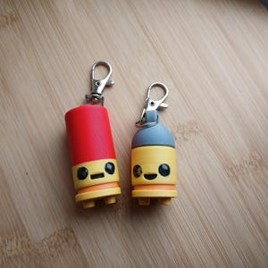 May include: Two novelty keychains with cartoon faces. One is red, the other yellow, both with black eyes and a small mouth. The keychains have silver clasps. The background is a light brown wooden surface.