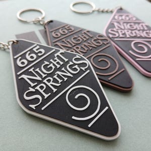 May include: Three keychains with a diamond-shaped tag. Each tag has the text "665 NIGHT SPRINGS" and a spiral design. The keychains are black, brown, and pink.