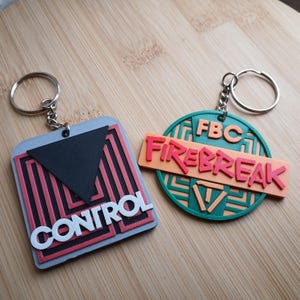 May include: Two keychains with 3D printed designs. One keychain is a square with a black triangle and the word "CONTROL" in white letters. The other keychain is a circle with a green and orange design and the words "FBC FIREBREAK" in red letters.