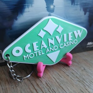Könnte beinhalten: Ein grüner und weißer Schlüsselanhänger in Form eines Motel-Schilds mit dem Text "Oceanview Motel and Casino".