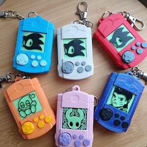 Puede incluir: Una colección de llaveros de consolas de juegos portátiles de colores. Los llaveros son de varios colores, incluyendo azul, blanco, rojo, naranja, rosa y morado. Cada llavero presenta un diseño de personaje diferente en la pantalla, con botones y un D-pad.