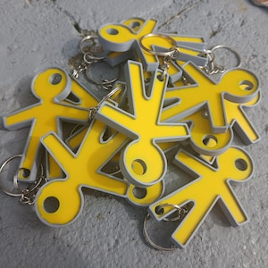 Op de afbeelding: Een verzameling gele sleutelhangers in de vorm van een stokfiguurtje met grijze randen. Elke sleutelhanger heeft een gestileerde menselijke vorm met een cirkelvormig hoofd en een gat voor een metalen ring. De figuren liggen over elkaar heen.