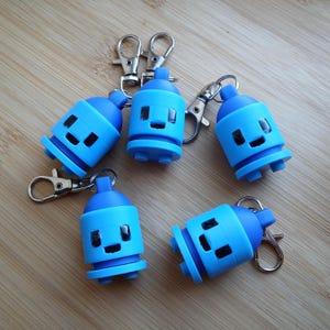 May include: Five blue spray paint can keychains with silver clasps. Each keychain has a smiling face design. The keychains are in various positions on a light brown wooden surface.