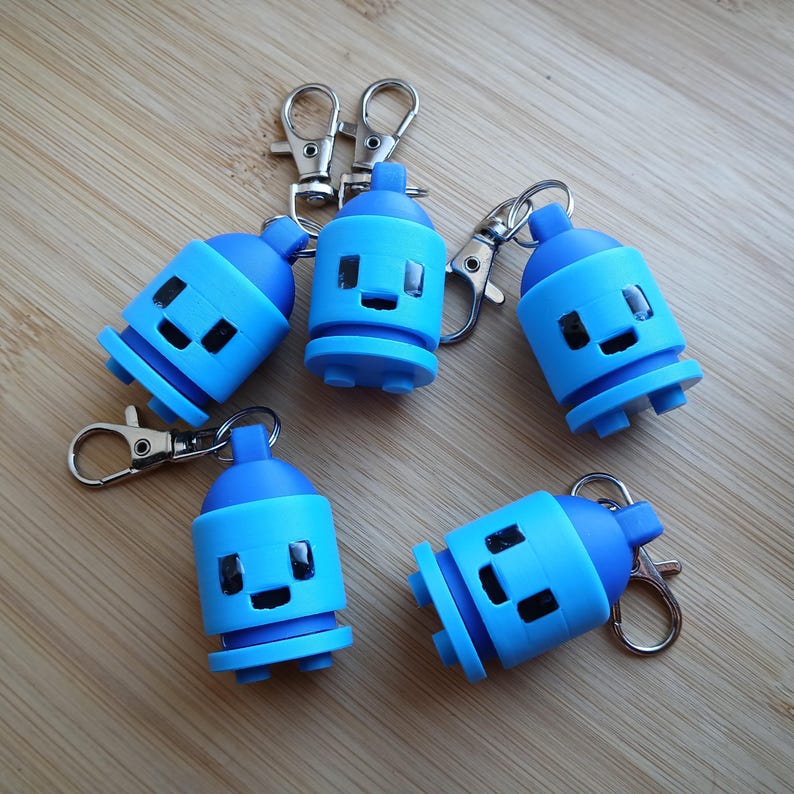 May include: Five blue spray paint can keychains with silver clasps. Each keychain has a smiling face design. The keychains are in various positions on a light brown wooden surface.