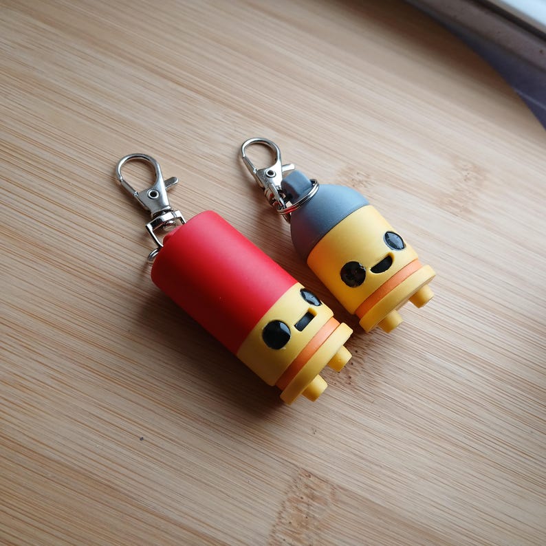 May include: Two novelty keychains with a cartoon design. One is red, the other is yellow and blue. Both have a silver clasp and a smiling face design. The keychains are on a wooden surface.