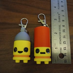 May include: Two novelty keychains with cartoon faces. One is yellow with a gray top, the other is yellow and orange. A ruler shows the keychains are approximately 2 inches (5 cm) tall. Both have silver-colored clasps.