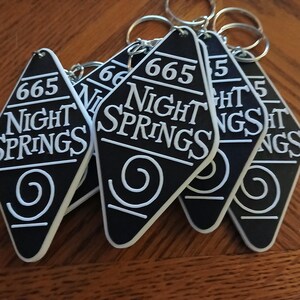 May include: Five black keychains shaped like motel room keys with white text that reads "665 Night Springs" and a white spiral design.