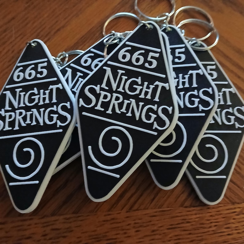 May include: Five black keychains shaped like motel room keys with white text that reads "665 Night Springs" and a white spiral design.