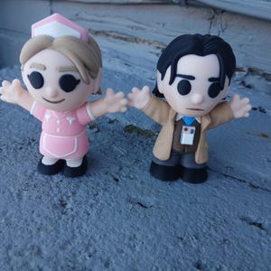 May include: Two small vinyl figurines standing side by side. The left figure is a person in a pink nurse uniform with a matching cap and blonde hair. The right figure is a person in a tan jacket, brown shirt, blue tie, and dark pants.