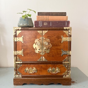 May include: A small, wooden cabinet with brass hardware and a drawer. The cabinet has a warm, reddish-brown finish and ornate brass accents. A small blue ceramic pot with a green plant and several vintage books are on top of the cabinet.