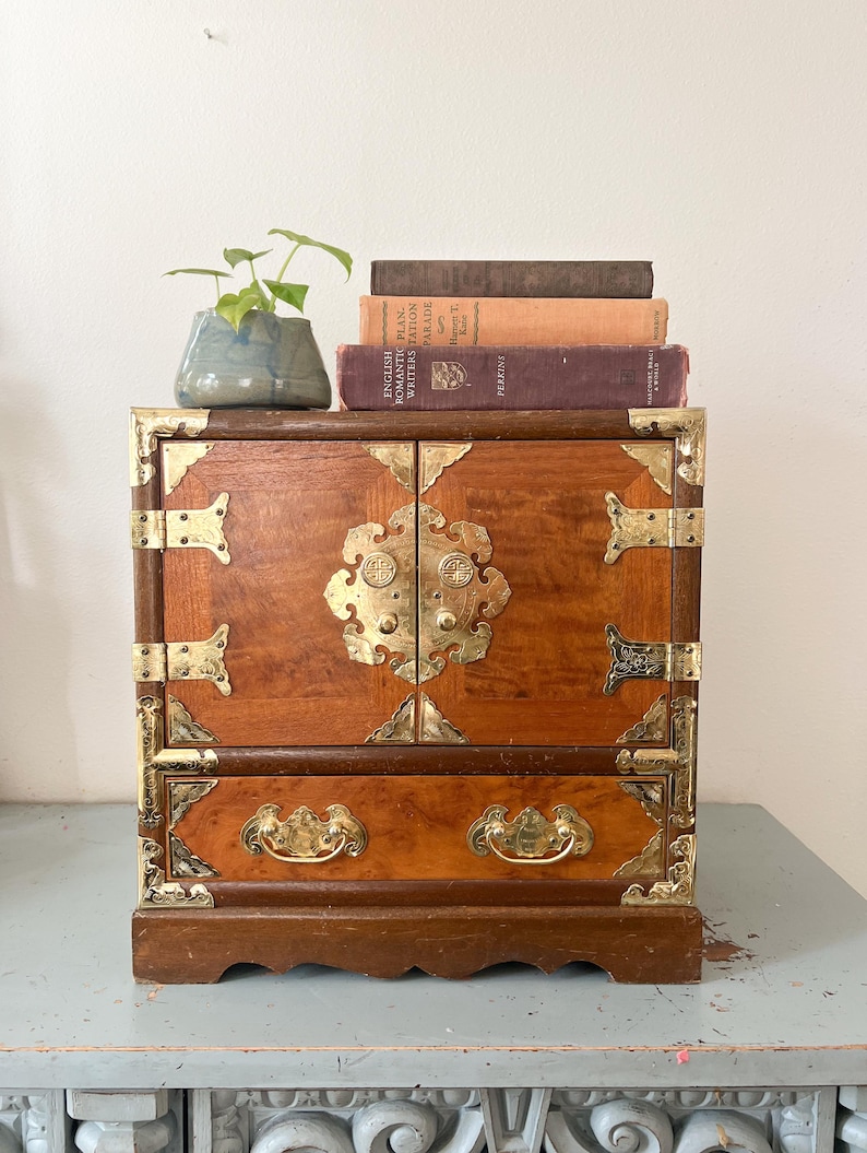 May include: A small, wooden cabinet with brass hardware and a drawer. The cabinet has a warm, reddish-brown finish and ornate brass accents. A small blue ceramic pot with a green plant and several vintage books are on top of the cabinet.