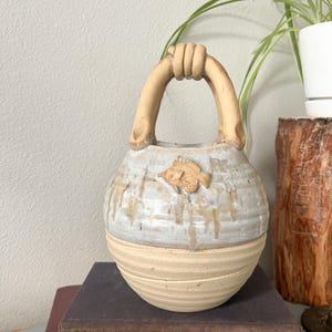 May include: A ceramic vase with a handle shaped like clasped hands. The vase is light beige with a white and beige glazed top and a fish-shaped embellishment. The vase is sitting on top of a stack of books.