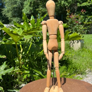 May include: A wooden articulated doll with jointed limbs, standing on a wooden base. The doll is made of light brown wood and has a simple, minimalist design.