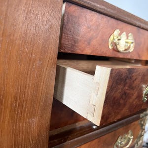 May include: A small, antique wooden chest of drawers with a warm, reddish-brown finish. The drawers have ornate brass handles and corner accents. One drawer is partially open, revealing the dovetail joinery.