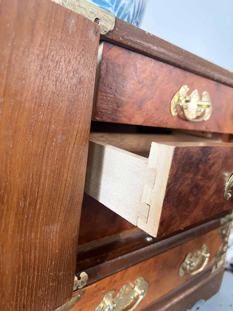 May include: A small, antique wooden chest of drawers with a warm, reddish-brown finish. The drawers have ornate brass handles and corner accents. One drawer is partially open, revealing the dovetail joinery.