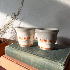 May include: Two off-white ceramic cups featuring a repeating pattern of orange, yellow, and white candy corn motifs around the center. Each cup has a flared rim and a thin orange horizontal band near the base, with a smooth, glossy finish.