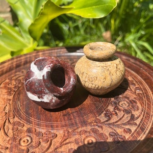 May include: Two small decorative pots, one is red and white marble with a hole in the side, the other is a light brown with a pattern, both are sitting on a carved wooden tray.