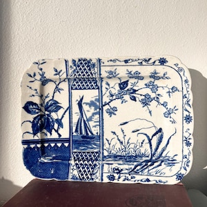 Könnte beinhalten: Rechteckige blaue und weiße Keramikservierplatte mit floralem und nautischem Design. Die Platte zeigt ein Segelboot, Blumen und Blätter. Die Kanten sind gewellt und mit floralen Mustern verziert.