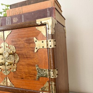 May include: A wooden chest with brass hardware, including corner brackets, hinges, and a decorative latch. The chest has a warm, reddish-brown finish and is topped with a stack of vintage books. The books have various colors and titles.