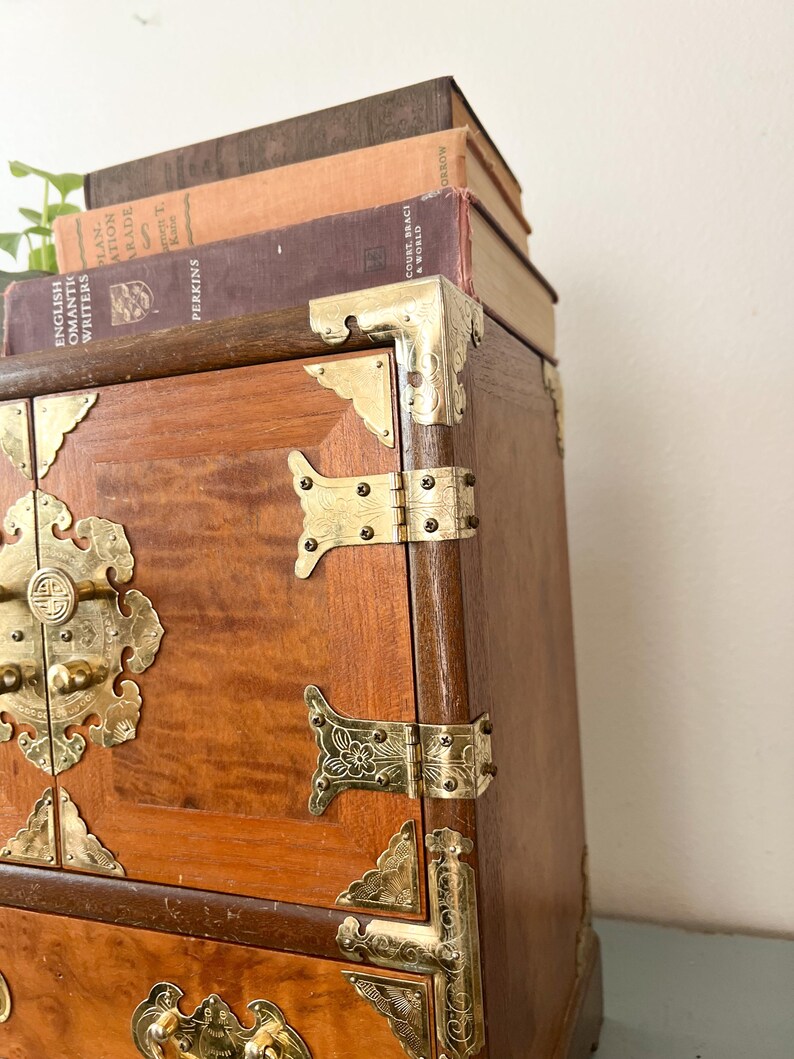 May include: A wooden chest with brass hardware, including corner brackets, hinges, and a decorative latch. The chest has a warm, reddish-brown finish and is topped with a stack of vintage books. The books have various colors and titles.