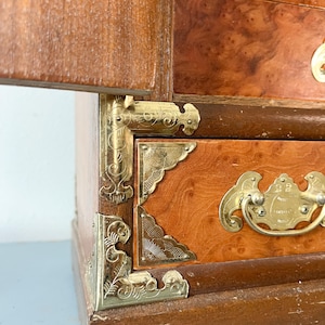 May include: A vintage wooden jewelry box with a warm, reddish-brown finish. The box features ornate brass corner brackets and drawer pulls. The wood grain is visible, and the box has a classic, antique aesthetic.