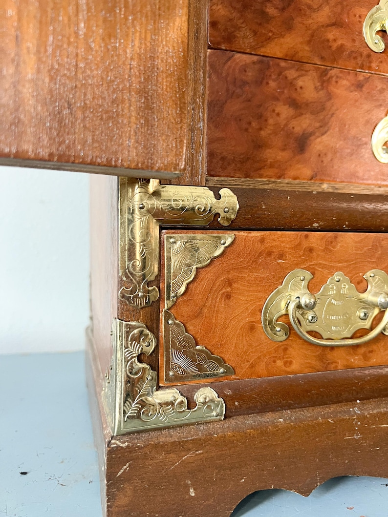 May include: A vintage wooden jewelry box with a warm, reddish-brown finish. The box features ornate brass corner brackets and drawer pulls. The wood grain is visible, and the box has a classic, antique aesthetic.