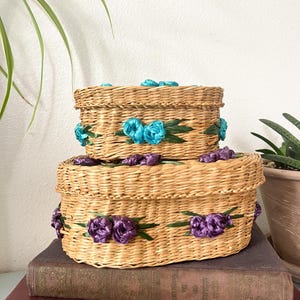 May include: Two woven wicker boxes, one stacked on top of the other. The boxes are decorated with teal and purple ribbon flowers and green leaves. The boxes are sitting on top of a stack of old books.
