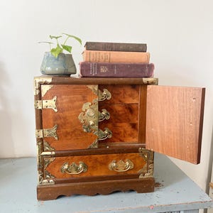 May include: A small, wooden jewelry box with brass hardware and a warm, reddish-brown finish. The box has multiple drawers and a hinged door, with decorative brass accents. A small potted plant and several books sit on top.