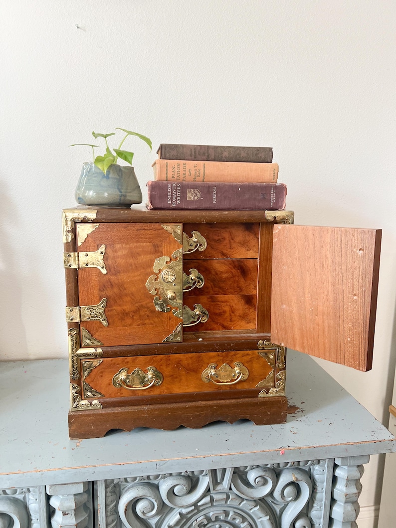 May include: A small, wooden jewelry box with brass hardware and a warm, reddish-brown finish. The box has multiple drawers and a hinged door, with decorative brass accents. A small potted plant and several books sit on top.