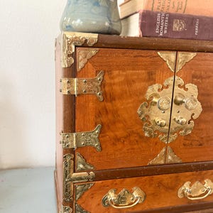 May include: A vintage wooden cabinet with brass hardware. The cabinet has a warm, reddish-brown finish and ornate brass corner brackets, hinges, and drawer pulls. A small blue ceramic pot with a green plant sits on top, along with a stack of old books.