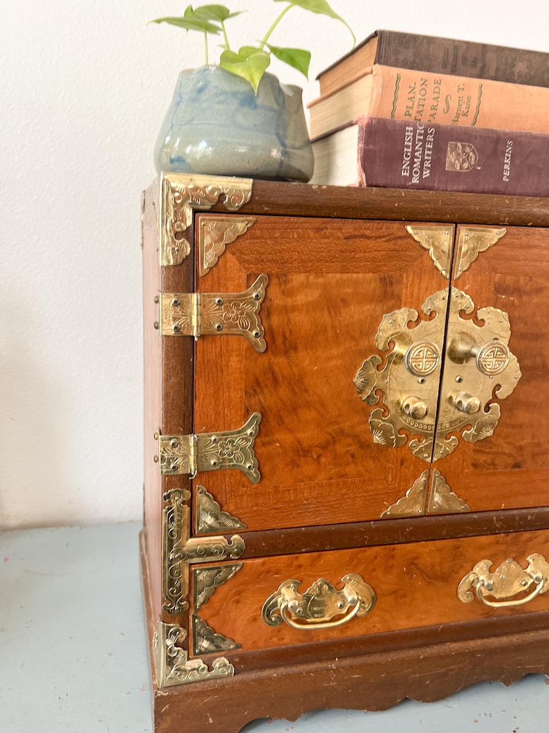 May include: A vintage wooden cabinet with brass hardware. The cabinet has a warm, reddish-brown finish and ornate brass corner brackets, hinges, and drawer pulls. A small blue ceramic pot with a green plant sits on top, along with a stack of old books.