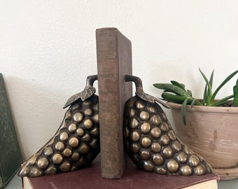 May include: Bronze grape cluster bookends with a vintage book between them. The bookends have a detailed texture and are placed on a maroon book. A potted plant is visible in the background.