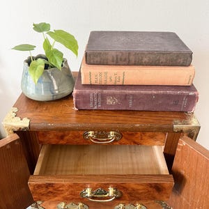 May include: A small, wooden nightstand with two drawers and brass hardware. The top of the nightstand holds a stack of vintage books and a small, blue-green ceramic pot with a green plant. The drawers are open, revealing the interior.