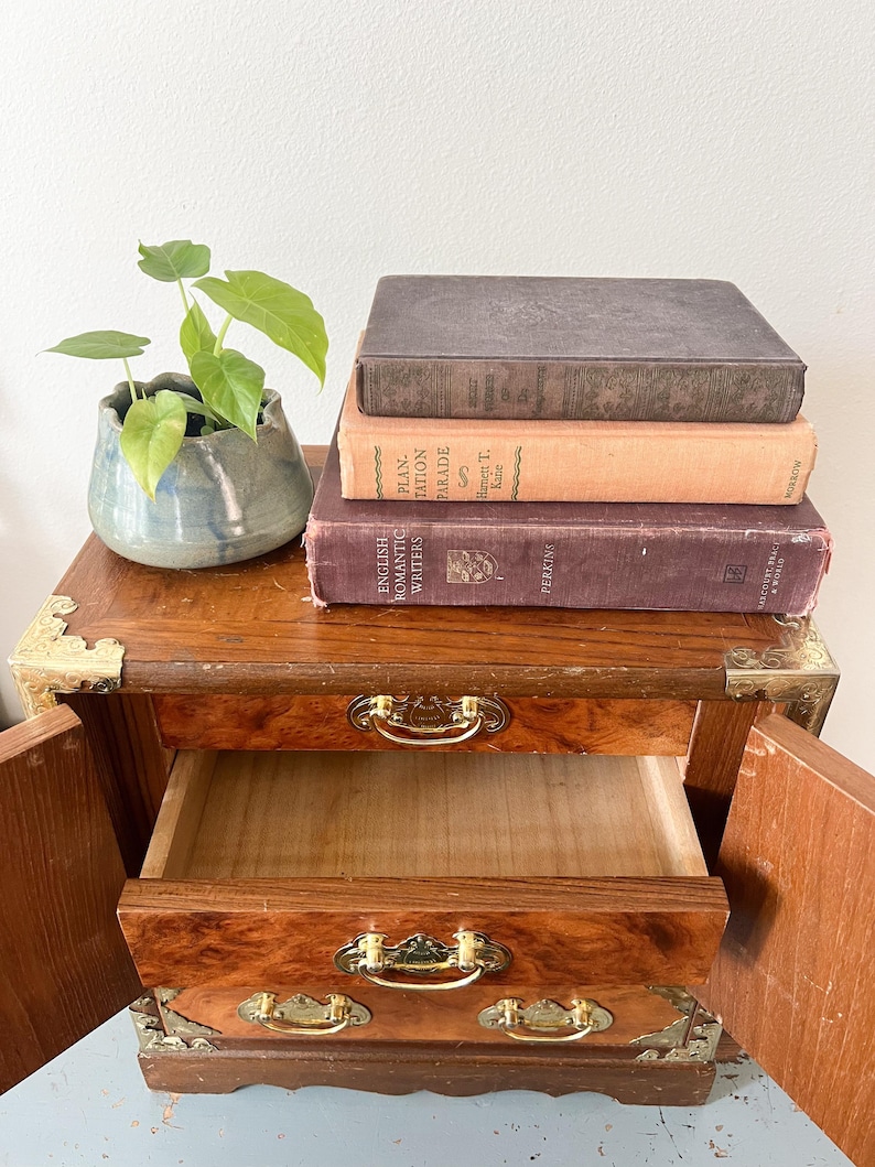 May include: A small, wooden nightstand with two drawers and brass hardware. The top of the nightstand holds a stack of vintage books and a small, blue-green ceramic pot with a green plant. The drawers are open, revealing the interior.