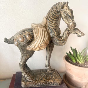 May include: A decorative horse figurine with a saddle, in a distressed black and gold finish. The horse is standing on a rectangular base, and the figurine is placed on top of two vintage books.