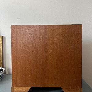 May include: A wooden cabinet with a warm brown finish. The cabinet has a rectangular shape with a base that is slightly lighter in color. The cabinet is set against a light-colored wall.