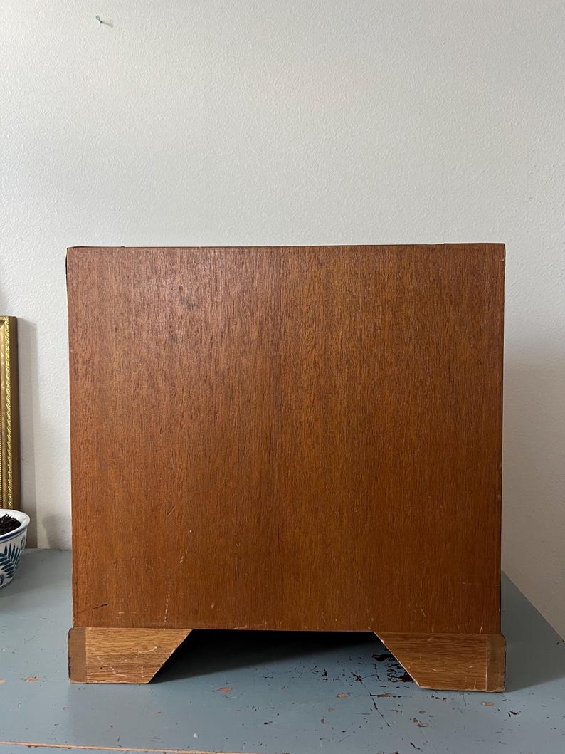 May include: A wooden cabinet with a warm brown finish. The cabinet has a rectangular shape with a base that is slightly lighter in color. The cabinet is set against a light-colored wall.