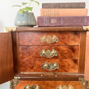 May include: A vintage wooden jewelry box with three drawers and brass hardware. The box has a burl wood finish and is open, revealing the drawers. A small potted plant and several antique books are on top.