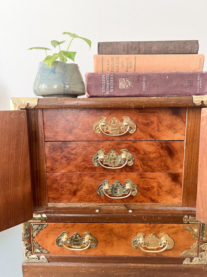 May include: A vintage wooden jewelry box with three drawers and brass hardware. The box has a burl wood finish and is open, revealing the drawers. A small potted plant and several antique books are on top.