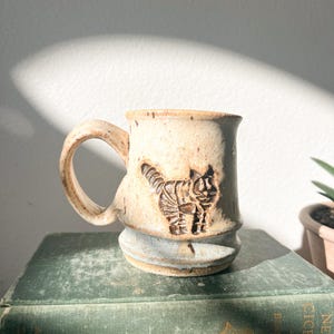 May include: A handmade ceramic mug with a cat design. The mug is beige with a brown cat illustration and a curved handle. The mug sits on a vintage green book. The mug is approximately 4 inches tall.